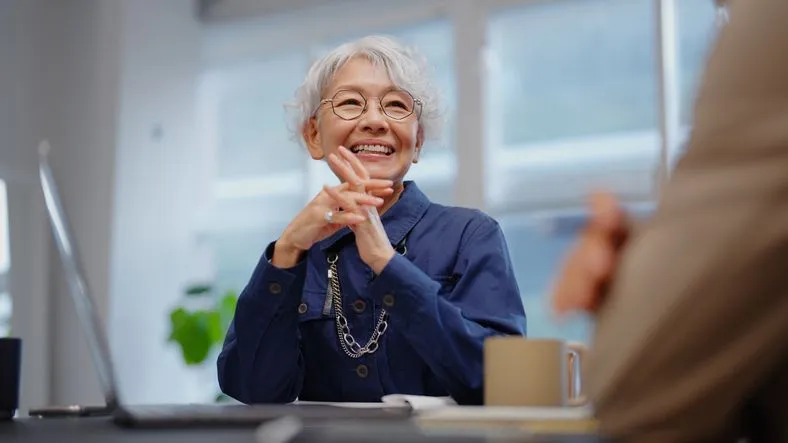 A professional older businesswoman in glasses and a navy blouse offers financial guidance to a senior businessman in a beige suit.