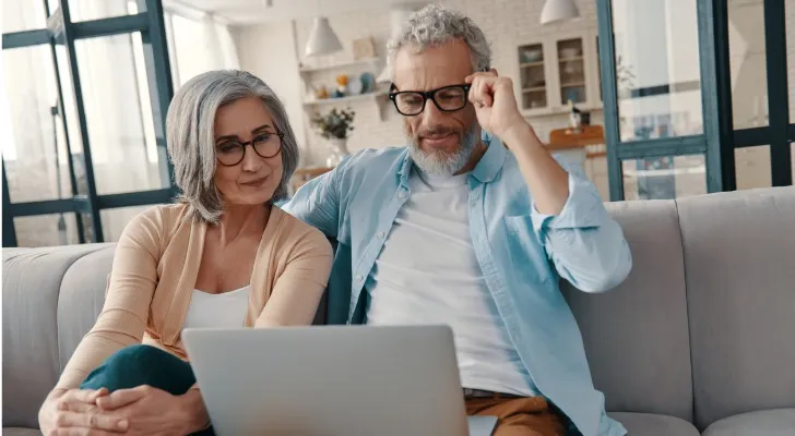 A husband and wife who appear to be in their 50s or 60s sit together on a couch and review information on a laptop.